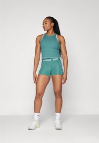 Teal treningssinglet og shorts med hvite detaljer og merkevare. Modellen står selvsikkert i hvite sneakers og sokker. Glatt stofftekstur.