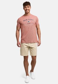 Homme souriant portant un t-shirt rouge avec un graphique New Port, un short beige et des baskets blanches, debout devant un fond clair uni.