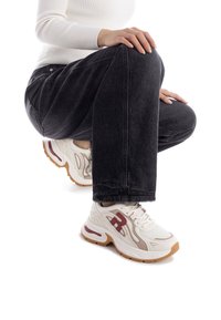 Sneaker bianca con dettagli rossi, upper in rete testurizzata, suola curva e ammortizzazione spessa. Indossate con jeans scuri in posizione accovacciata.