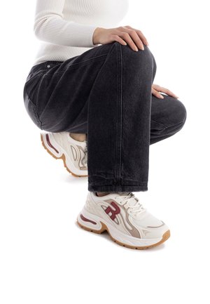 Zapatilla blanca con detalles rojos, parte superior de malla texturizada, suela curva y un grueso acolchado. Llevada con vaqueros oscuros en una posición agachada.