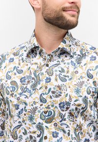 Heren overhemd met knopen, met een witte basis en een kleurrijk paisley- en bloemenpatroon in blauw, groen, geel en bruin.