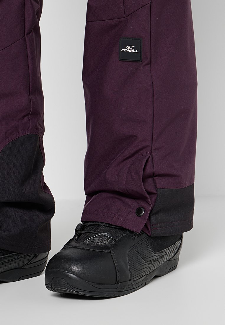 Fioletowe spodnie narciarskie O'Neill zakrywają czarne buty snowboardowe z zapiętymi mankietami i widocznym logo marki na nogawce spodni.