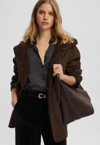 Cappotto marrone con colletto in shearling, camicia grigia con bottoni, pantaloni di velluto a coste neri, fibbia dorata e borsa scura in mano.