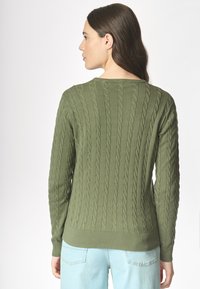 Una donna con i capelli lunghi indossa un maglione a trecce verde scuro, girata di spalle, abbinato a jeans azzurri su uno sfondo bianco semplice.