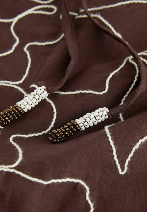 Brauner Stoff mit weißem Stickmuster. Baumwollkordeln sind an den Enden mit kleinen weißen und braunen Perlen für zusätzliche Details versehen.