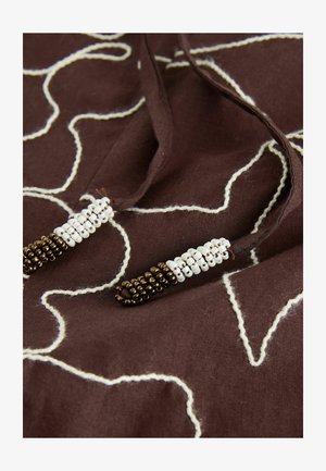 Tissu marron avec motif brodé blanc. Les cordons en coton sont ornés de petites perles blanches et marron aux extrémités pour un détail supplémentaire.
