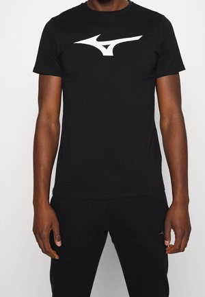 Person trägt ein eng anliegendes schwarzes Kurzarmshirt mit großem weißem abstraktem Logo auf der Brust, kombiniert mit schwarzen Hosen.