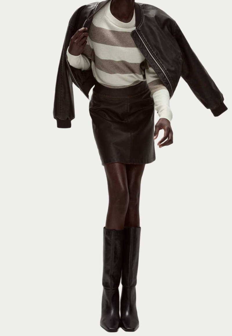 Personne portant un pull rayé beige et blanc, une jupe en cuir noire, de hautes bottes noires, tenant une veste en cuir noire sur les épaules.