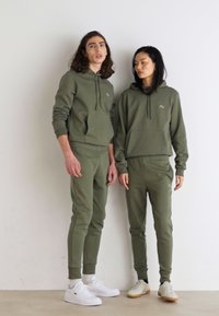 Olivgrüne Kapuzenpullover und passende Jogginghosen aus weichem Material, mit einem kleinen Logo auf der Brust. Getragen mit weißen und beigen Sneakers.
