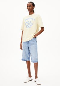 Gelbes Übergröße-T-Shirt mit blauem Blumenmotiv und Schriftzug, kombiniert mit hellblauen Jeansshorts und weißen Schnürschuhen.