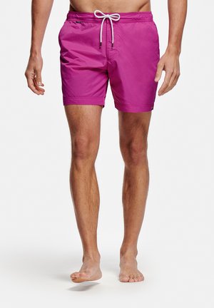 Man draagt felroze zwembroek met witte trekkoord, staat op blote voeten tegen een witte achtergrond.