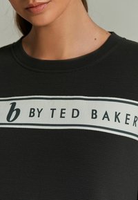 Czarny sweter z dzianiny o prążkowanej teksturze, z białym paskiem z napisem "b BY TED BAKER" w czarnym kolorze.