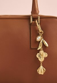 Sac à main en cuir marron avec un charme doré orné de motifs floraux et de feuilles, incluant un accent en perle, attaché à la fermeture éclair.
