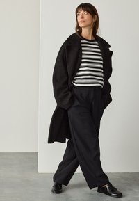 Cappotto nero con texture sopra una camicia a righe bianco e nero, abbinato a pantaloni neri larghi e scarpe nere lucide. Design semplice e monocromatico.