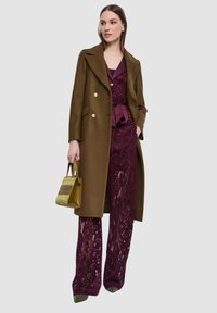 Cappotto doppiopetto marrone, tuta in pizzo color granata, borsa verde, tacchi a punta. La tuta ha pantaloni ampi e una cintura in raso.