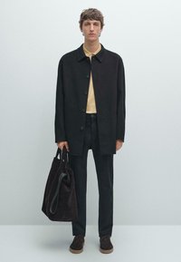 Manteau oversize noir, polo jaune, pantalon noir, sac en suede marron et chaussures brun foncé. Texture lisse, détails de boutons, design minimaliste.