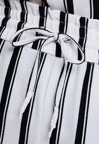 Tissu rayé noir et blanc avec une finition texturée, doté d'une taille à cordon de serrage et de liens blancs. Les lignes verticales créent un motif défini.
