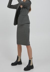 Kobieta ubrana w czarno-białą kratkę marynarkę i spódnicę, czarny top oraz czarno-beżowe masywne botki na kostkach na jednolitym tle.