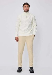 Pull blanc en maille texturée avec col zippé, motifs en losange, associé à un pantalon beige ajusté et des chaussures noires. Le mannequin est debout, face, sur un fond neutre.