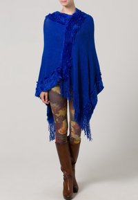 Poncho blu lavorato a maglia con bordi frangiati e bordo testurizzato. Indossato sopra leggings colorati con motivi e stivali marroni sopra il ginocchio.