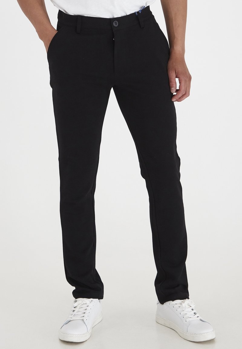 Sorte slim-fit bukser med en glat tekstur, der har en knaplukning, sidelommer og diskrete syninger, parret med hvide sneakers.