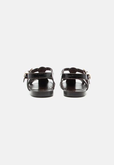 Μαύρα γυαλιστερά σανδάλια με επίπεδη σόλα, λουριά στο πίσω μέρος και κούμπωμα με χρυσή πόρπη, εμφανιζόμενα από πίσω σε απλό λευκό φόντο.