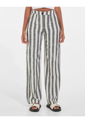 Nainen, jolla on päällään korkeavyötäröiset, leveälahkeiset housut, joissa on pystysuorat mustavalkoiset raidat ja beiget solkisandaalit valkoisella taustalla.