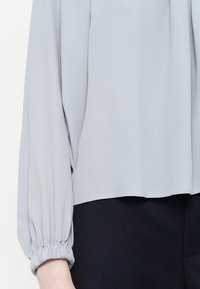 Helle graue Bluse aus glattem Stoff, mit langen, gerafften Ärmeln und lockerer Passform, kombiniert mit dunklen Hosen.