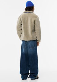 Moški v bež flis jakni, širokih modrih kavbojkah, svetlo modri kapi in rjavih čevljih, stoji z obrnjeno hrbtno stranjo.