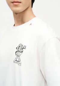 Κοντινό πλάνο ατόμου που φορά άσπρο πουκάμισο με μαύρο γραφικό Moka pot γεμάτο λουλούδια και κείμενο «Breakfast Klub» στο στήθος.