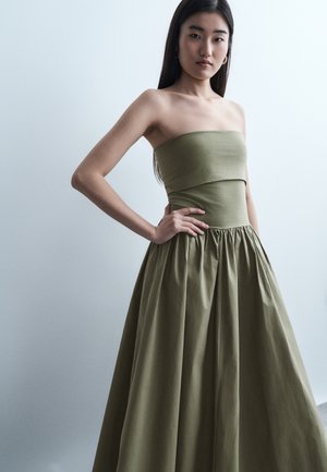 Νέα γυναίκα στέκεται με το χέρι στη μέση φορώντας φόρεμα χωρίς τιράντες σε ελιά πράσινο χρώμα με πτυχωτή φούστα, μπροστά σε απλό ανοιχτό φόντο.