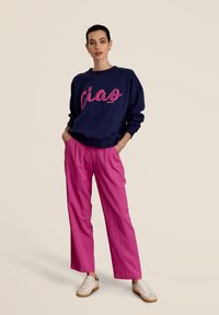 Marineblauwe sweatshirt met roze "ciao" tekst en geribbelde manchetten, gecombineerd met felroze wijd-uitlopende broeken en crèmekleurige sneakers op een lichte achtergrond.