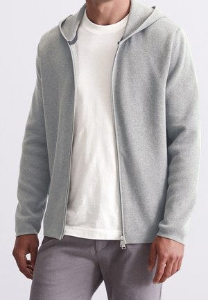 Mann trägt hellgrauen Kapuzenpullover mit Reißverschluss über weißem T-Shirt und grauer Hose, steht vor einfarbigem Hintergrund.