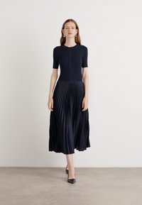 Navy blue dress with a fitted top and pleated, flowy skirt. Short sleeves, textured fabric, and a knee-length hem. Worn with black heels.