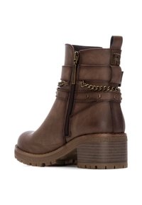Botín de cuero marrón con cremallera lateral, detalle de cadena y un tacón de bloque texturizado. Presenta tachuelas decorativas y un refuerzo en la parte trasera.