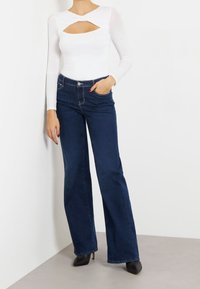 Woman wearing a white long-sleeve top with a chest cutout and dark blue high-waisted wide-leg jeans, standing with one hand in pocket.