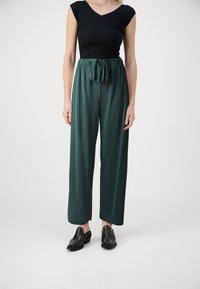 Pantalons teal à jambes larges avec une taille à cordon, associés à un haut noir ajusté. Modèle portant des chaussures noires à enfiler, se tenant devant un fond clair.