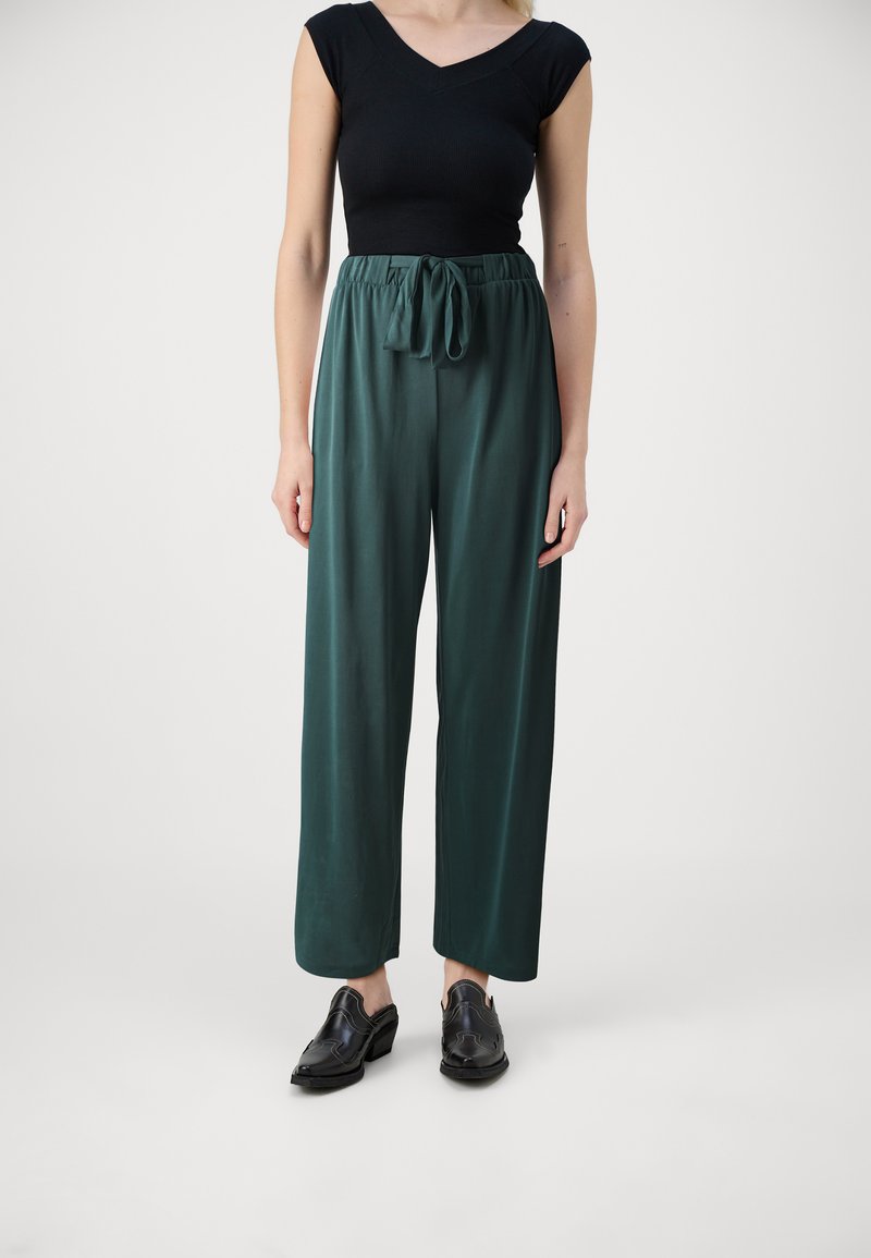 Pantalons teal à jambes larges avec une taille à cordon, associés à un haut noir ajusté. Modèle portant des chaussures noires à enfiler, se tenant devant un fond clair.