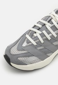Gray athletic shoe with mesh and suede materials, featuring white laces, textured patterns, and a black rubber outsole.