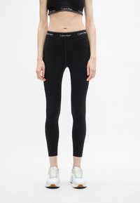 Čierne legíny s priliehavým strihom a elastickým materiálom, so širokým pásom s logom Calvin Klein. V kombinácii s bielymi teniskami.