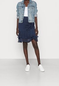 Jeansjacka med ljus tvätt, långa ärmar, framfickor, över en marinblå prickig volangkjol. Modellen bär vita sneakers.