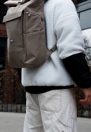 Person trägt eine weiße Fleecejacke und beige Cargohosen, mit einem taupefarbenen rechteckigen Rucksack über einer Schulter im Freien.