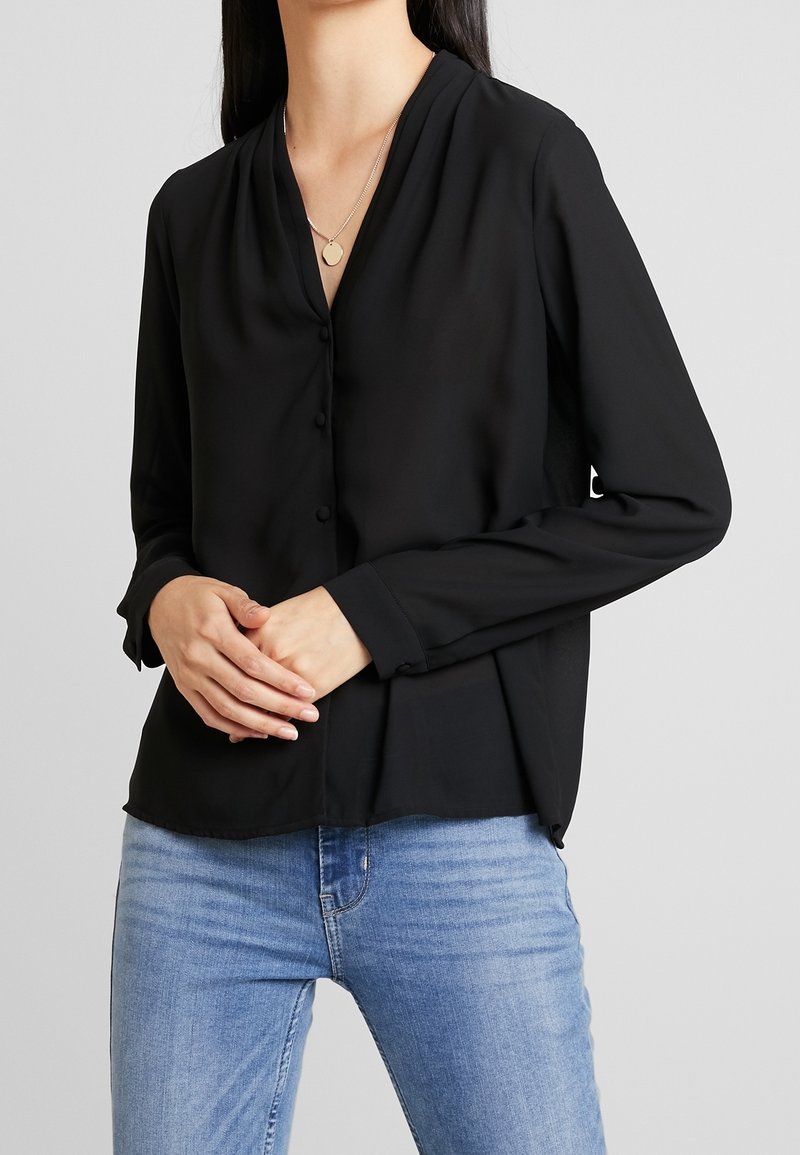 Femme portant un chemisier noir à manches longues avec un col en V et des boutons, ainsi qu'un jean bleu clair, les mains jointes devant, sur un fond neutre.