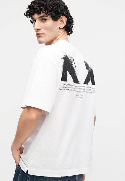 Άνδρας με σγουρά μαλλιά που φοράει λευκό φαρδύ μπλουζάκι με μαύρο γραφικό και κείμενο στην πλάτη, συνδυασμένο με σκούρο μπλε τζιν.