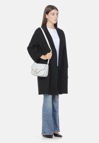 Cappotto nero oversize, t-shirt bianca, jeans blu a zampa d'elefante e scarpe nere appuntite. Porta una borsa grigia chiara con una chiusura dorata.