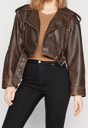 Osoba ubrana w skróconą brązową skórzaną kurtkę z detalami zamka błyskawicznego, na brązowej bluzce i czarnych spodniach z wysokim stanem.