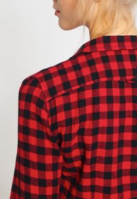 Röd och svart rutig flanellskjorta med krage, långa ärmar och rundad nederkant. Mjuk textur med en strukturerad design.