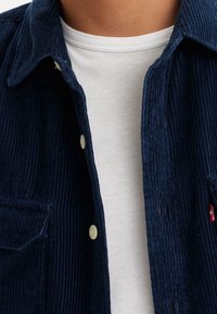 Marinblå manchester-skjorta med knäppning fram, två bröstfickor och en nedvikt krage, lager-på-lager över en vit t-shirt med rund halsringning.