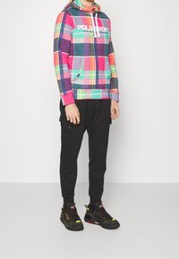 Felpa a scacchi colorata nei toni del rosa, verde e viola, con tasca a marsupio e logo bianco; abbinata a pantaloni cargo neri e sneaker nere.