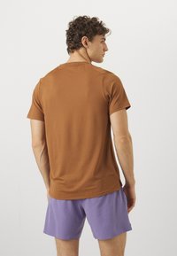 T-shirt castanho de mangas curtas feito de um tecido suave, com decote redondo e fendas laterais, usado com calções roxos.